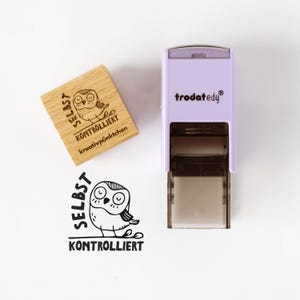 Könnte beinhalten: Ein Holzstempel mit einer Eulenillustration und den deutschen Wörtern "SELBST KONTROLLIERT" und "kreativpünktchen". Ein lila-weißer Stempel mit dem Markennamen "trodat edy" ist ebenfalls zu sehen. Das Eulenmotiv wiederholt sich darunter.