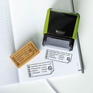 Könnte beinhalten: Ein grüner und schwarzer Trodat printy 4913 Stempel neben einem Holzstempel und gestempelten Checklisten auf einem Notizbuch. Die Checklisten umfassen Schulmaterialien wie Papier, Stifte und Kleber. Der Holzstempel hat das gleiche Checklisten-Design.