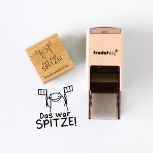 Op de afbeelding: Een houten stempel en een zelfinktende stempel met de tekst "Das war SPITZE!" en een stripfiguur met vlaggen. De houten stempel heeft de tekst "kreativpünktchen". De zelfinktende stempel is beige en zwart.