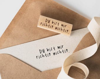 Può includere: Una busta marrone con un biglietto bianco all'interno. Il biglietto ha il testo "DU bist mir richtig wichtig." scritto su di esso. Un timbro di legno con lo stesso testo è sopra la busta.