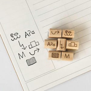 May include: Wooden stamps with various symbols and letters, including "A?a", "M", "~", "<->", "&uarr;", "&darr;", and a book. The stamps are arranged in a cluster on a white notebook page.