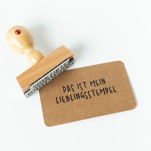 May include: A wooden stamp with a red top and the text "DAS IST MEIN LIEBLINGSSTEMPEL" on a brown card. The stamp is made of light-colored wood and has a rectangular base with a metal stamping surface.