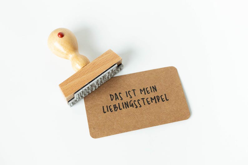 May include: A wooden stamp with a red top and the text "DAS IST MEIN LIEBLINGSSTEMPEL" on a brown card. The stamp is made of light-colored wood and has a rectangular base with a metal stamping surface.