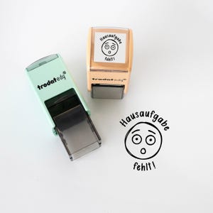 Könnte beinhalten: Zwei Stempel, einer mintgrün und einer beige, mit dem Text "Hausaufgabe fehlt!" und einem überraschten Gesicht. Der mintgrüne Stempel trägt den Markennamen "trodat edy".