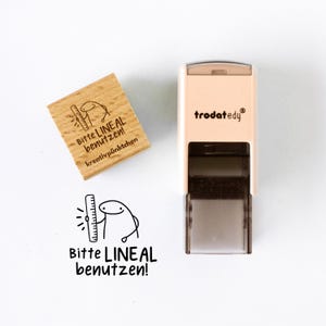 Könnte beinhalten: Ein Holzstempel und ein Selbstfärbestempel mit dem Text "Bitte LINEAL benutzen!" und einer Comicfigur, die ein Lineal hält. Der Holzstempel trägt den Text "kreativpünktchen". Der Selbstfärbestempel ist beige und trägt den Markennamen "trodat edy".