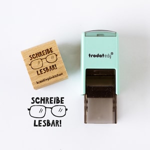 May include: A wooden stamp with the words "SCHREIBE LESBAR!" and a glasses graphic. A mint green stamp with the text "trodat edy" is also in the image. The stamped image of the glasses and text is below the wooden stamp.