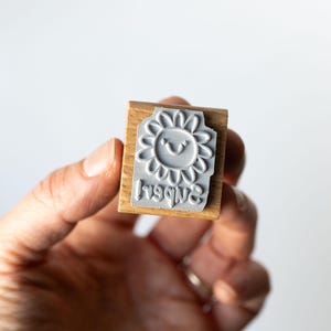 May include: A wooden stamp with a gray rubber stamp of a smiling sunflower and the word "happy" in reverse. The stamp is held in a hand against a white background. The stamp is a craft supply.