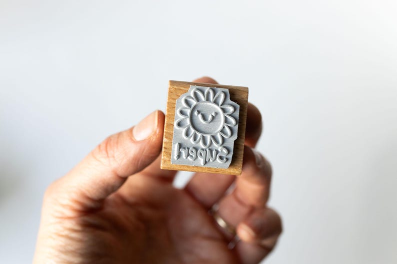 May include: A wooden stamp with a gray rubber stamp of a smiling sunflower and the word "happy" in reverse. The stamp is held in a hand against a white background. The stamp is a craft supply.