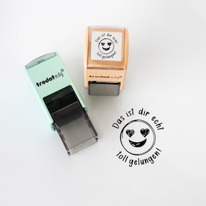 Könnte beinhalten: Zwei Selbstfärbestempel, einer mintgrün und einer beige, mit dem Text "Das ist dir echt toll gelungen!" um ein Smiley-Gesicht mit Herzaugen. Die Stempel befinden sich auf einer weißen Oberfläche.