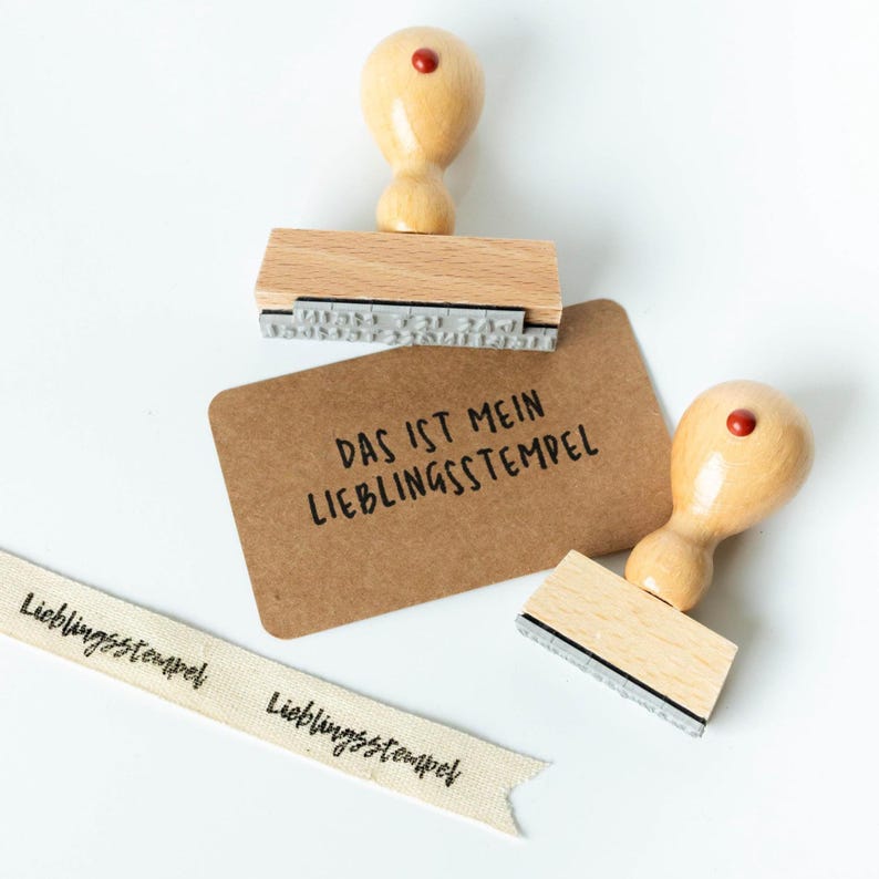 May include: Wooden stamps with a light brown handle and a red dot on top. The stamps are next to a brown card with the text "DAS IST MEIN LIEBLINGSSTEMPEL" and a ribbon with the text "Lieblingsstempel".