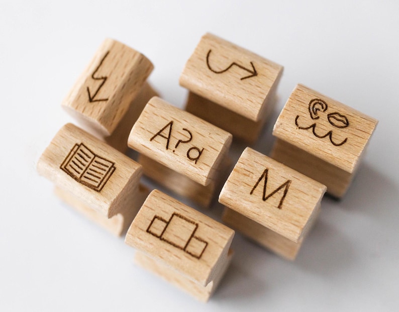 May include: Wooden stamps with various symbols and letters, including an arrow, a book, a wave, lips, a letter M, and the letters A?a. The stamps are made of light brown wood and have a natural finish.