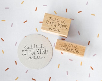 Può includere: Tre timbri in legno e un adesivo rotondo con la scritta "Endlich SCHULKIND - Mathilda" in carattere scritto a mano. I timbri sono rettangolari e l'adesivo è beige. Lo sfondo è bianco con coriandoli sparsi.