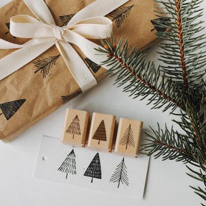 Peut inclure: Un ensemble de quatre tampons en bois repr&eacute;sentant un motif d'arbre simple. Les tampons sont dispos&eacute;s en rang&eacute;e sur une surface blanche, avec un morceau de papier sur lequel des arbres sont tamponn&eacute;s en dessous. Un sac cadeau brun avec un ruban blanc et une branche de pin sont &eacute;galement pr&eacute;sents sur l'image.