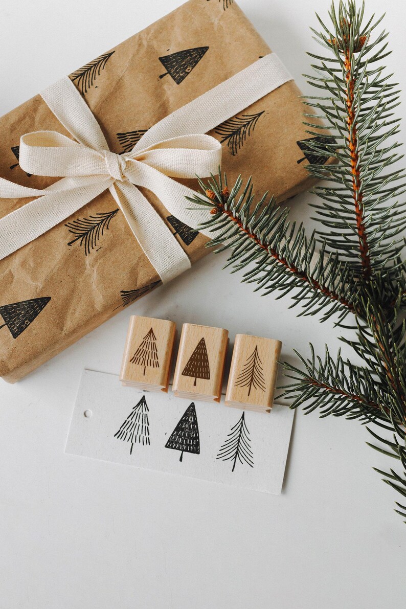 Peut inclure: Un ensemble de quatre tampons en bois repr&eacute;sentant un motif d'arbre simple. Les tampons sont dispos&eacute;s en rang&eacute;e sur une surface blanche, avec un morceau de papier sur lequel des arbres sont tamponn&eacute;s en dessous. Un sac cadeau brun avec un ruban blanc et une branche de pin sont &eacute;galement pr&eacute;sents sur l'image.