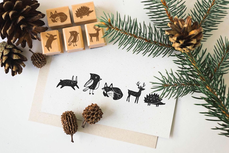 K&ouml;nnte beinhalten: Holzstempel mit Waldtier-Designs, darunter Hase, Vogel, Fuchs, Hirsch und Igel. Die Stempel sind mit Tannenzapfen und Tannenzweigen angeordnet, was ein rustikales, von der Natur inspiriertes Thema schafft.