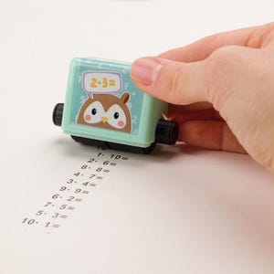 May include: A mint green rolling stamp featuring an owl cartoon and the equation "2+3=". The stamp is rolling across a white surface, leaving a series of math problems. Black wheels and a black handle are visible.