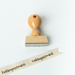 May include: A wooden stamp with a rounded handle and a red dot on top. The stamp's base is rectangular, and the text "Lieblingsstempel" is visible on a ribbon. The ribbon is beige with black text.
