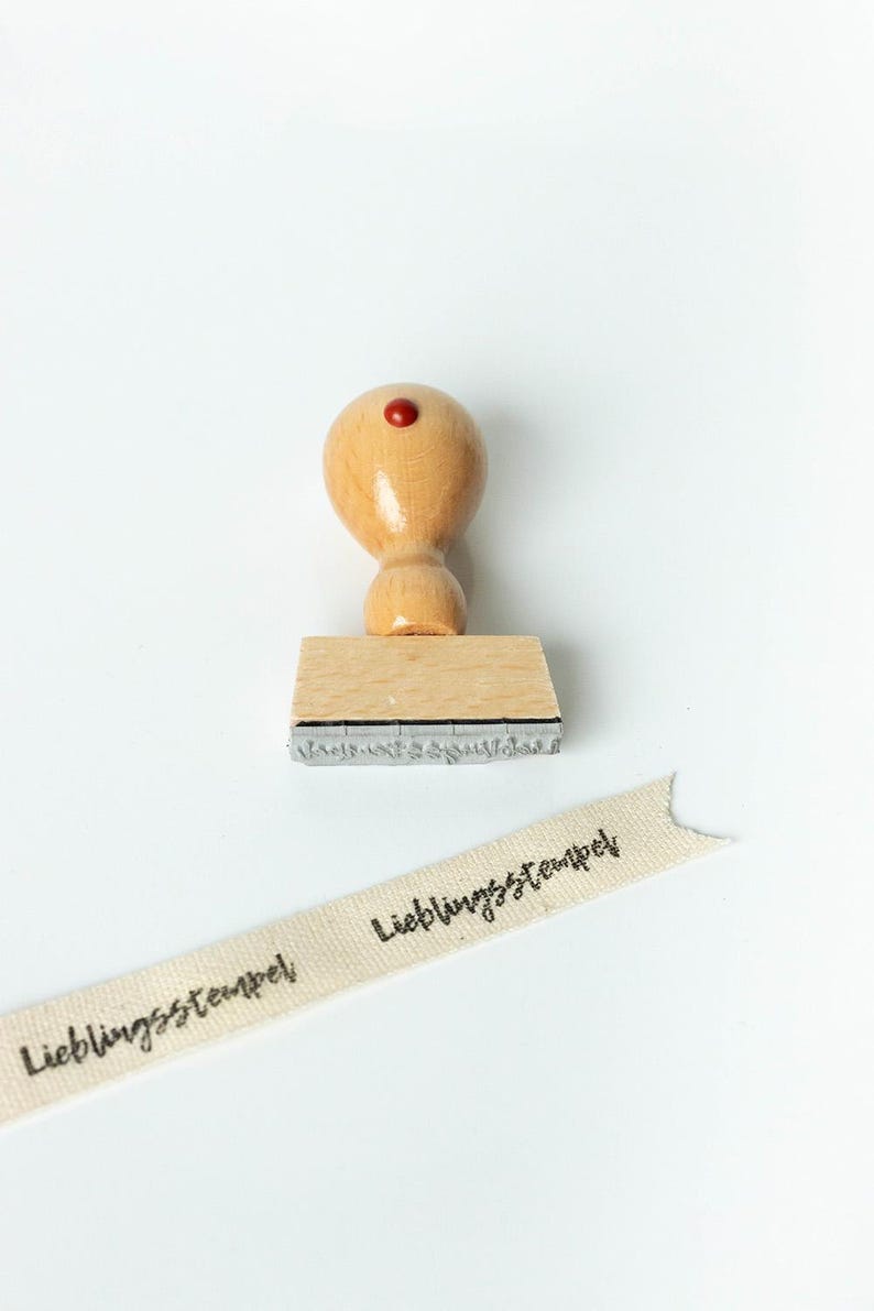 May include: A wooden stamp with a rounded handle and a red dot on top. The stamp's base is rectangular, and the text "Lieblingsstempel" is visible on a ribbon. The ribbon is beige with black text.