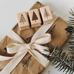 Peut inclure: Un sac cadeau en papier brun avec un ruban blanc et trois tampons en bois avec un motif d'arbre. Le sac est d&eacute;cor&eacute; d'un motif d'arbres r&eacute;p&eacute;t&eacute;. Une branche de pin est en arri&egrave;re-plan.