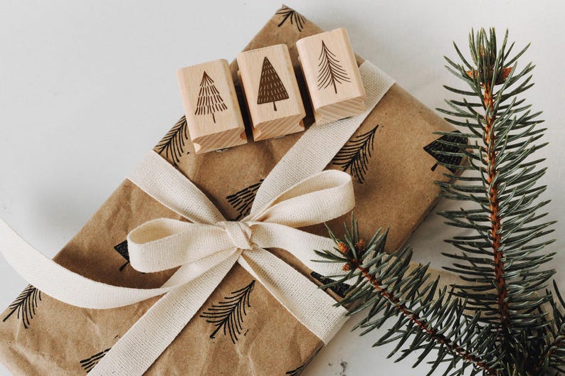 Peut inclure: Un sac cadeau en papier brun avec un ruban blanc et trois tampons en bois avec un motif d'arbre. Le sac est d&eacute;cor&eacute; d'un motif d'arbres r&eacute;p&eacute;t&eacute;. Une branche de pin est en arri&egrave;re-plan.