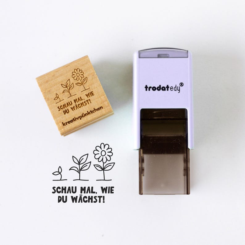 Op de afbeelding: Een houten stempel met de tekst "SCHAU MAL, WIE DU W&Auml;CHST!" en een bloemenontwerp, naast een witte en zwarte stempel met de merknaam "trodatedy". De afbeelding toont ook de gestempelde afbeelding van het bloemenontwerp en de tekst.