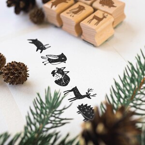 K&ouml;nnte beinhalten: Holzstempel mit Tiermotiven, darunter ein Reh und ein Fuchs, sind auf einer wei&szlig;en Oberfl&auml;che angeordnet. Die Stempel haben eine nat&uuml;rliche Holzoberfl&auml;che. Schwarze Tintenabdr&uuml;cke der Tiere sind auf wei&szlig;em Papier gedruckt. Tannenzapfen und gr&uuml;ne Zweige sind ebenfalls vorhanden.