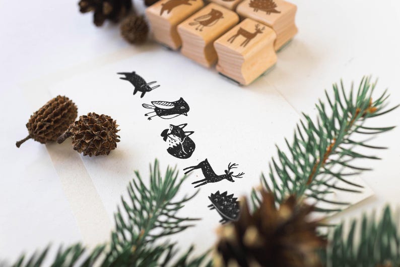 K&ouml;nnte beinhalten: Holzstempel mit Tiermotiven, darunter ein Reh und ein Fuchs, sind auf einer wei&szlig;en Oberfl&auml;che angeordnet. Die Stempel haben eine nat&uuml;rliche Holzoberfl&auml;che. Schwarze Tintenabdr&uuml;cke der Tiere sind auf wei&szlig;em Papier gedruckt. Tannenzapfen und gr&uuml;ne Zweige sind ebenfalls vorhanden.