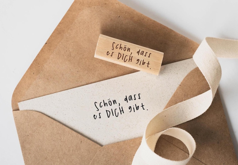 May include: A brown paper envelope with a white card inside. The card has the text "Sch&ouml;n, dass es DICH gibt." written on it. A wooden stamp with the same text is on top of the envelope.