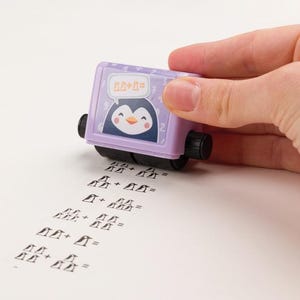 Könnte beinhalten: Ein lila Rollstempel mit einem Pinguin-Motiv und dem Text "2+2=". Der Stempel rollt über eine weiße Oberfläche und hinterlässt eine Spur von Pinguinen und mathematischen Symbolen. Der Stempel wird von einer Hand gehalten.