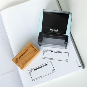Könnte beinhalten: Ein hellblau-schwarzer Trodat printy 4913 Stempel und ein Holzstempel mit dem Text "INFO FÜR DIE ELTERN". Das Bild zeigt auch zwei gedruckte Vorlagen mit dem gleichen Text und eine Bleistiftillustration.