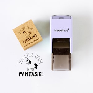 May include: A wooden stamp with the text "ICH LIEBE DEINE FANTASIE!" and a unicorn illustration. A white and black stamp with the brand name "trodat edy" is also in the image. The stamp is designed for creative projects.