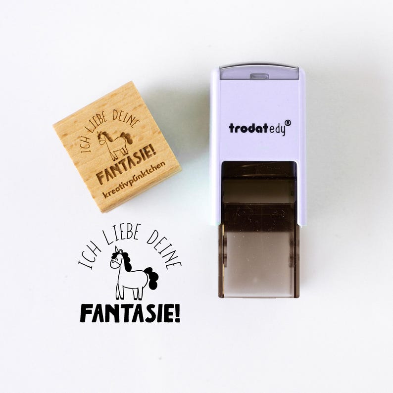 May include: A wooden stamp with the text "ICH LIEBE DEINE FANTASIE!" and a unicorn illustration. A white and black stamp with the brand name "trodat edy" is also in the image. The stamp is designed for creative projects.