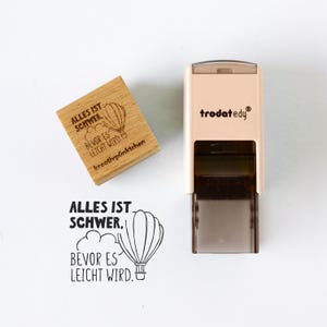 Può includere: Un timbro in legno con il testo tedesco "ALLES IST SCHWER, BEVOR ES LEICHT WIRD" e un'illustrazione di mongolfiera. È presente anche un secondo timbro con lo stesso testo e la stessa illustrazione, insieme a un timbro beige con il marchio "trodatedy".