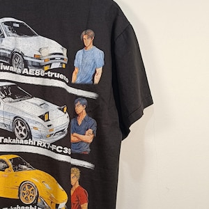 Könnte beinhalten: Ein schwarzes Kurzarm-T-Shirt mit einem Grafikdruck von drei Sportwagen und drei Charakteren. Der Text lautet Initial D, AE86-trueno, Takahashi RX7-FC3S und Takahashi RX7-FD3S. Die Autos sind weiß, weiß und gelb.