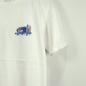 May include: A white short-sleeve t-shirt featuring a graphic on the left chest. The graphic displays a blue wave design, two surfboards, and the text HAWAII U.S.A. in blue block letters. The shirt has a crew neckline and a standard fit.