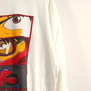May include: A white long-sleeve t-shirt featuring a vertical graphic print on the left side. The print consists of three stacked panels depicting anime-style characters, including a close-up of eyes, a character wearing goggles, and a red motorcycle.