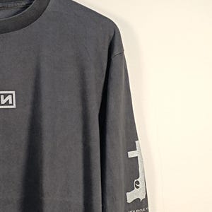 以下が含まれることがあります： 胸元に白いボックスロゴ、袖に白い拳銃のグラフィックがプリントされたチャコールグレーの長袖Tシャツ。袖には「NINE INCH NAILS」の文字と、NINの文字が入った正方形のロゴがデザインされています。