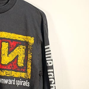 May include: A charcoal gray long-sleeve t-shirt featuring a distressed yellow and red square graphic on the chest with the text downward spirals below it. The sleeve displays the white text nine inch na in a vertical orientation along the arm.