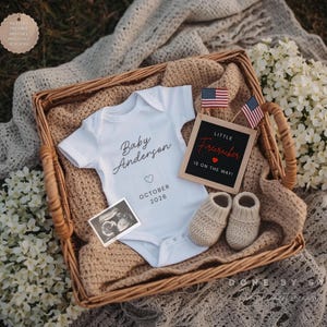 May include: A wicker basket holds a white baby onesie with "Baby Anderson" printed on it, a sonogram photo, and a letter board that reads "Little Firecracker is on the way!" with two small American flags. Beige knitted baby booties complete the arrangement.