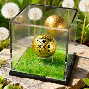 May include: A gold golf ball with the text "FORE-EVER S G 06.13.26" inside a clear display case with artificial grass. A second gold golf ball is visible in the background. The case sits on a rock surface with dandelions in the background.