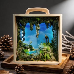 Pode incluir: Cena de diorama em miniatura dentro de uma caixa de madeira com uma alça. A cena retrata uma caverna com uma figura segurando uma luz, uma pequena piscina de água e um fundo de céu azul. A caixa está em uma bandeja de madeira.