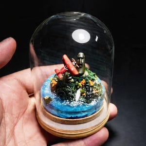 May include: A miniature diorama of a tropical island scene, featuring a red biplane and a Moai statue, encased in a glass dome. The island has lush green vegetation, orange flowers, and blue water. The base is made of wood.