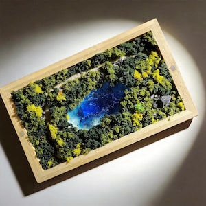 Puede incluir: Diorama de paisaje en miniatura con un lago azul vibrante rodeado de exuberante follaje verde y amarillo. La escena está encerrada en un marco de madera de color claro, creando un efecto tridimensional. Un pequeño sendero serpentea por el bosque.