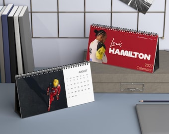 Puede incluir: Un calendario de escritorio 2027 con Lewis Hamilton. La portada es roja con texto blanco y una foto del piloto. La página abierta muestra el mes de agosto con una fotografía de un piloto con traje de carreras rojo y casco amarillo en la pista.