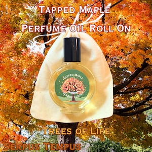 May include: A perfume oil roll-on bottle with a black cap and a label featuring a tree design. The bottle is inside a cream-colored pouch. The text "TAPPED MAPLE" and "PERFUME OIL ROLL ON" are visible. The background shows autumn foliage.