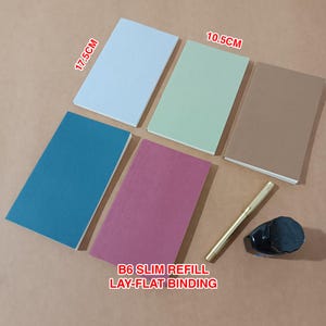 May include: A selection of B6 slim refill notebooks in shades of blue, green, and pink, featuring a lay-flat binding. The notebooks measure approximately 17.5cm by 10.5cm. A gold pen and a black ink bottle are also included in the image.