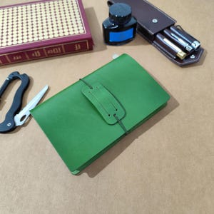 May include: A rectangular green leather journal secured with a matching green elastic band and a central leather tab. The journal is positioned alongside a red hardcover book, a black ink bottle, a brown leather pen case with two pens, and a black carabiner.
