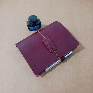 May include: A deep burgundy leather journal with a vertical elastic strap closure and a matching leather tab. A silver fountain pen is tucked under the strap, and a small glass bottle of blue ink with a black cap sits next to the journal.