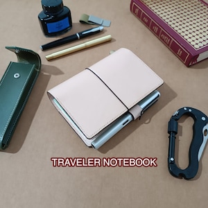 May include: A light beige leather traveler notebook with a black elastic band, holding a silver pen. Surrounding items include a green leather case, a black ink bottle, a black and gold pen, a brass pen, a red book, and a black carabiner tool.