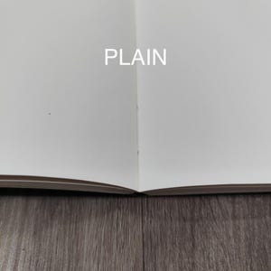 以下が含まれることがあります： 空白の白い無地のページが開かれたノート。中央の折り目には綴じ糸が見える。2ページの中央には白い大文字で「PLAIN」という文字が印刷されている。紙は滑らかでマットな質感である。
